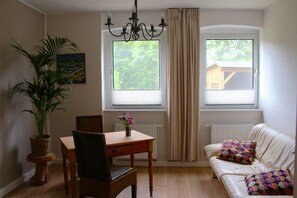 A room with a dining table, chairs, a potted plant, a sofa with patterned cushions, and a window with curtains. - Vacation apartment 1 - Green idyll (Borrentin)
