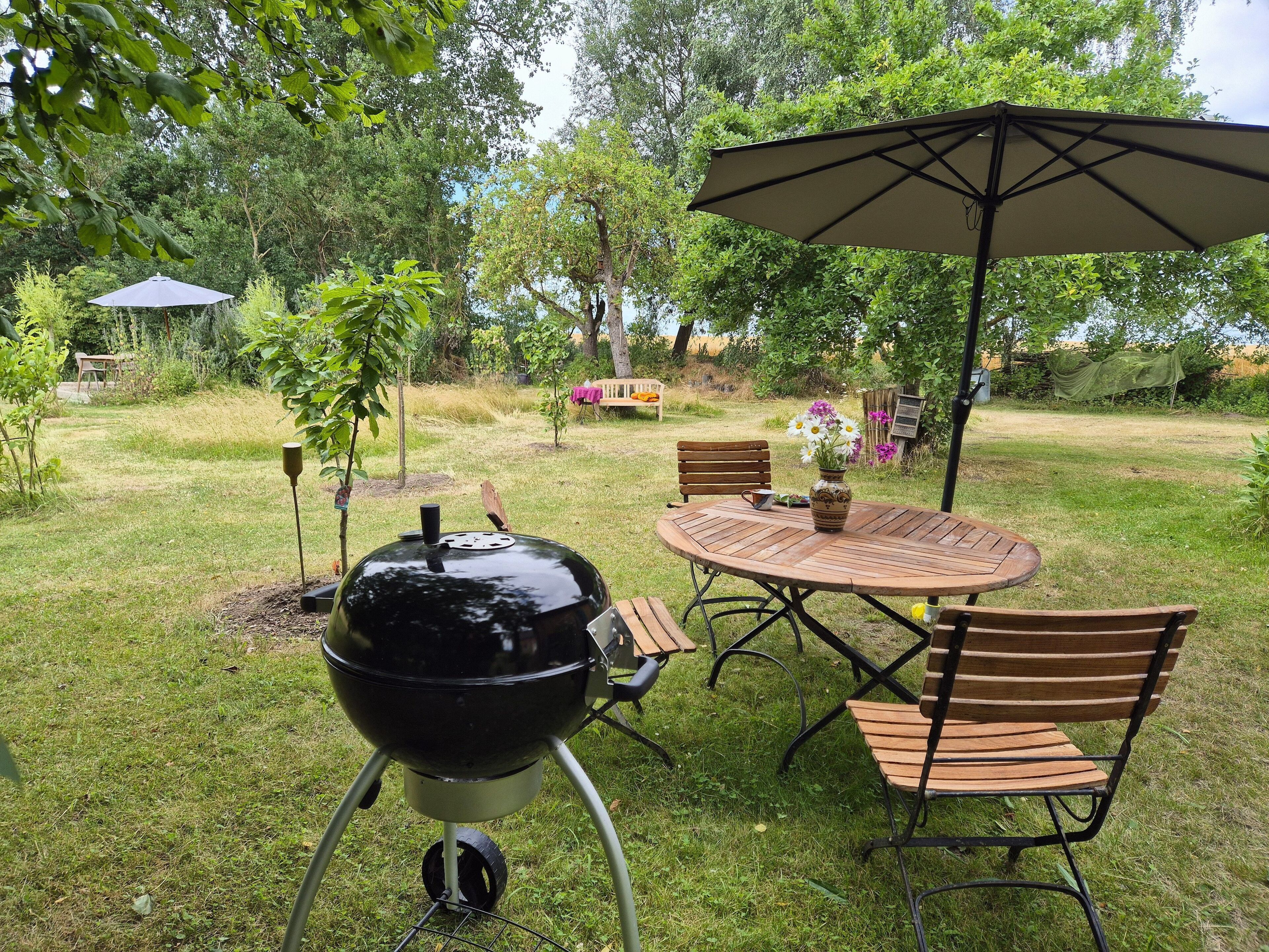 En trädgård med en grill, ett runt träbord, två trästolar och ett stort parasoll.