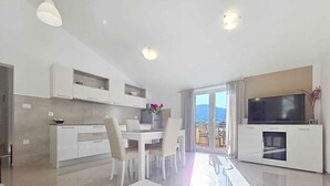 Cocina moderna con armarios blancos, comedor con mesa y sillas blancas, balcón con televisor y un ventanal grande.