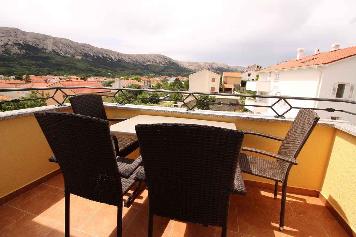 A balcony with wicker chairs and a table, overlooking a town with red-roofed buildings and mountains in the background.