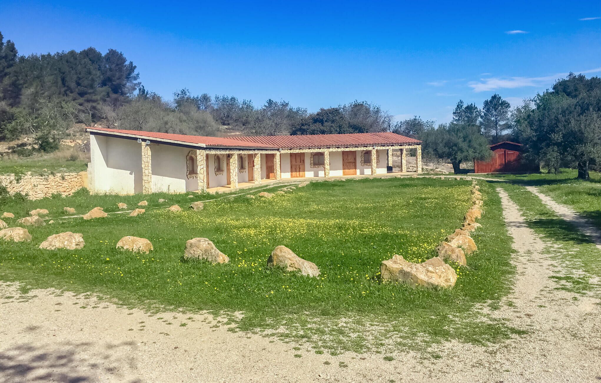 Un edificio de piedra con techo de tejas rojas, un porche cubierto y un sendero de tierra que lleva a él.