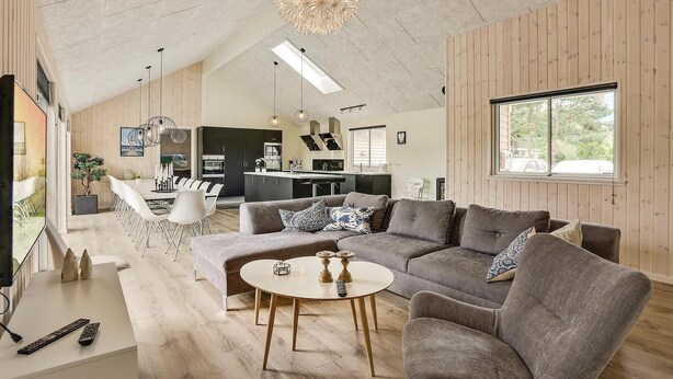 A spacious living room with a grey sectional sofa, a round coffee table, and a dining area with white chairs. A kitchen is visible in the background with modern appliances.