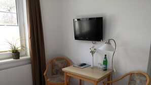 Una stanza piccola con un tavolo in legno e due sedie, una TV montata a parete, una lampada, una pianta in vaso e una bottiglia d'acqua.