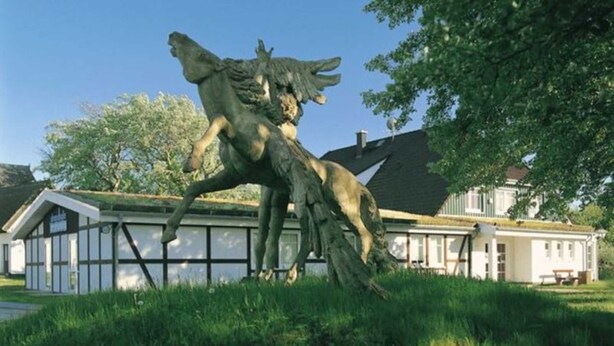 Una scultura che raffigura un lupo e un cervo in una zona erbosa, con una casa sullo sfondo.