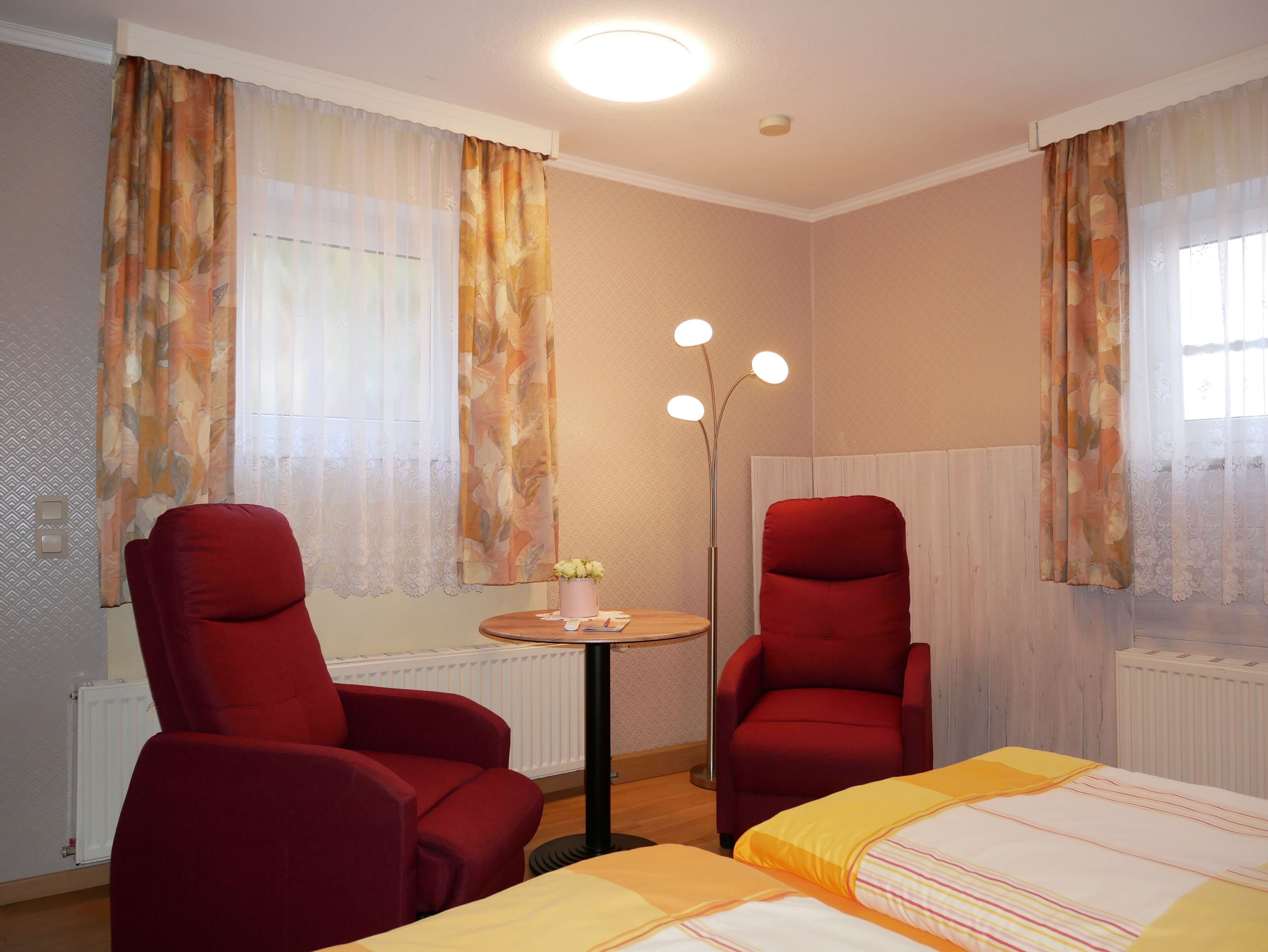A hotel room with two red chairs, a bed with a striped bedspread, a small round table with a vase of flowers, and a tall floor lamp.