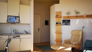 Un espace de vie compact comprenant un coin cuisine, une chaise, un canapé et une petite étagère agrémentée d’objets décoratifs.