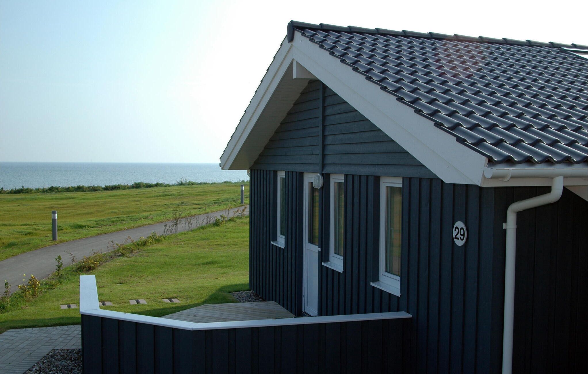 Een klein, donker gebouw met een schuin dak en een bord met het nummer '29', gelegen nabij een grasveld met een pad dat naar de zee leidt.