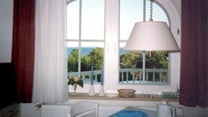 Quarto com uma grande janela, um radiador branco, uma cadeira branca, uma mesa com um tabuleiro azul e um abajur branco.