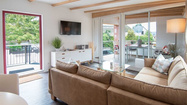 Un salón moderno con sofá, televisor, una mesa de centro de vidrio y un balcón con vistas a zonas verdes.