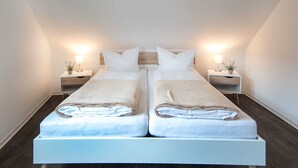 Una cama con dos plazas y cabecera de madera, dos mesitas de noche con lámparas y una pequeña planta en cada una.