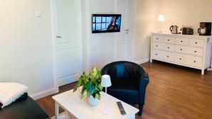 Un salon comprenant un téléviseur, un canapé, une table basse avec une plante, et un coin cuisine avec une machine à café.