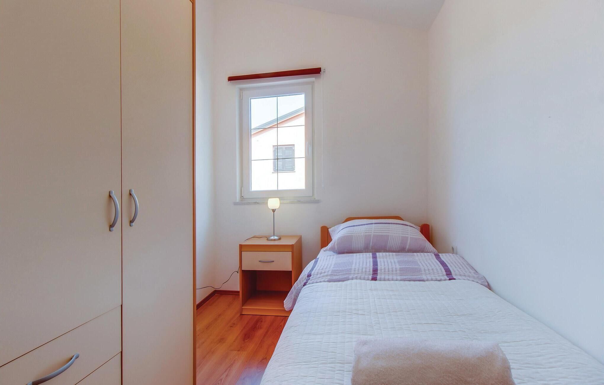 Ein kleines, gut beleuchtetes Schlafzimmer mit einem Einzelbett, einem Nachttisch, einer Nachttischlampe und einem Fenster mit Jalousien.