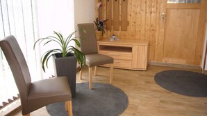 Een kamer met een houten bureau, twee stoelen, een potplant en een groot raam met jaloezieën.