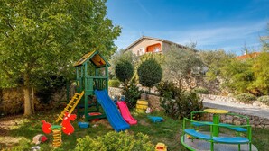Un parque infantil con tobogán, escalera de cuerda y columpios, rodeado de vegetación y un muro de piedra.