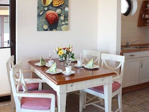 Ein Essbereich mit einem Holztisch, gedeckt für vier Personen, weißen Stühlen mit rosafarbenen Kissen und einem Bild von Meeresschnecken an der Wand.