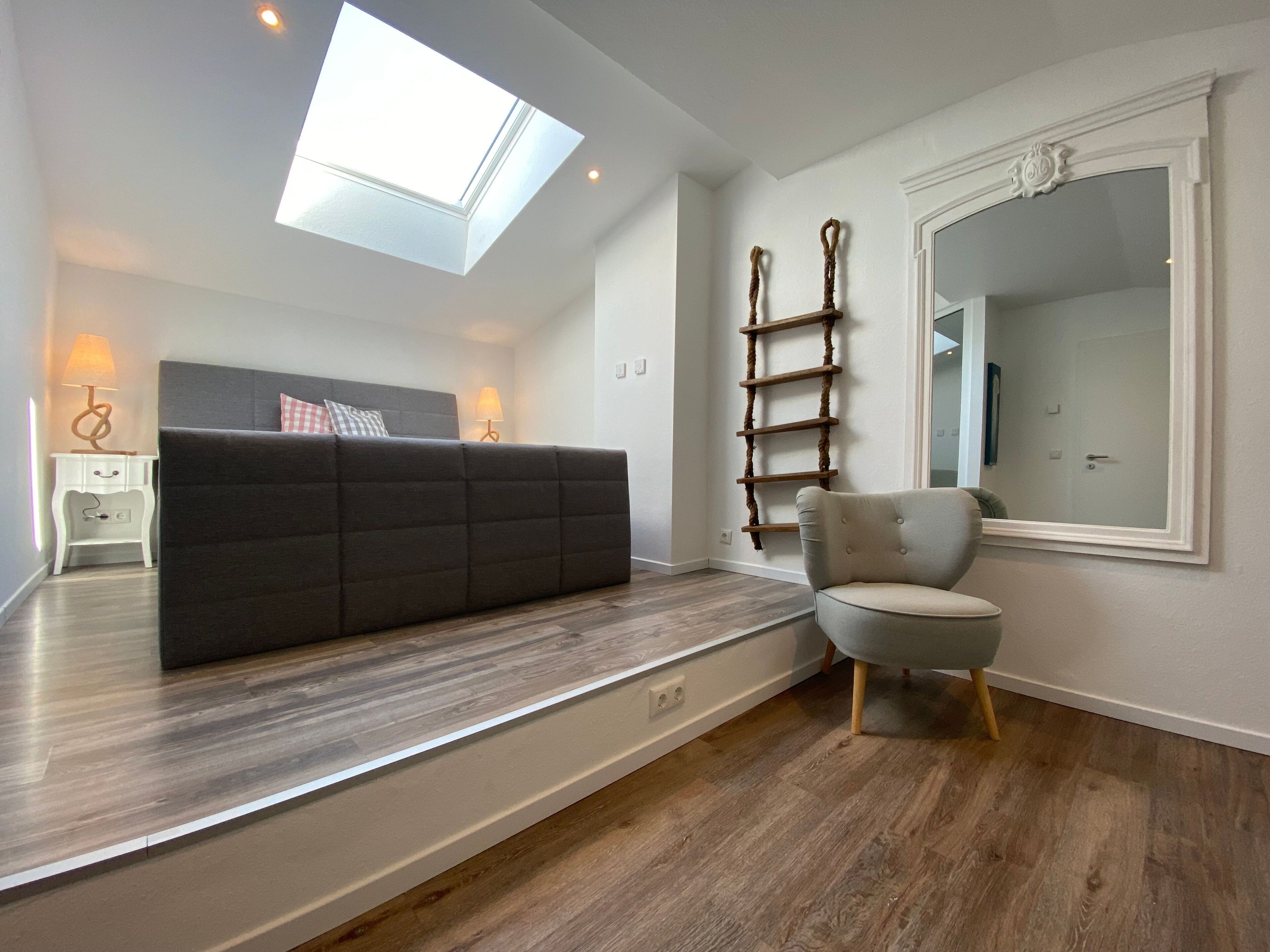 Een moderne woonkamer met een donkere bank, een lichte fauteuil, een houten laddenplank en een grote spiegel aan de muur.