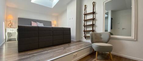 Een moderne woonkamer met een donkere bank, een lichte fauteuil, een houten laddenplank en een grote spiegel aan de muur.