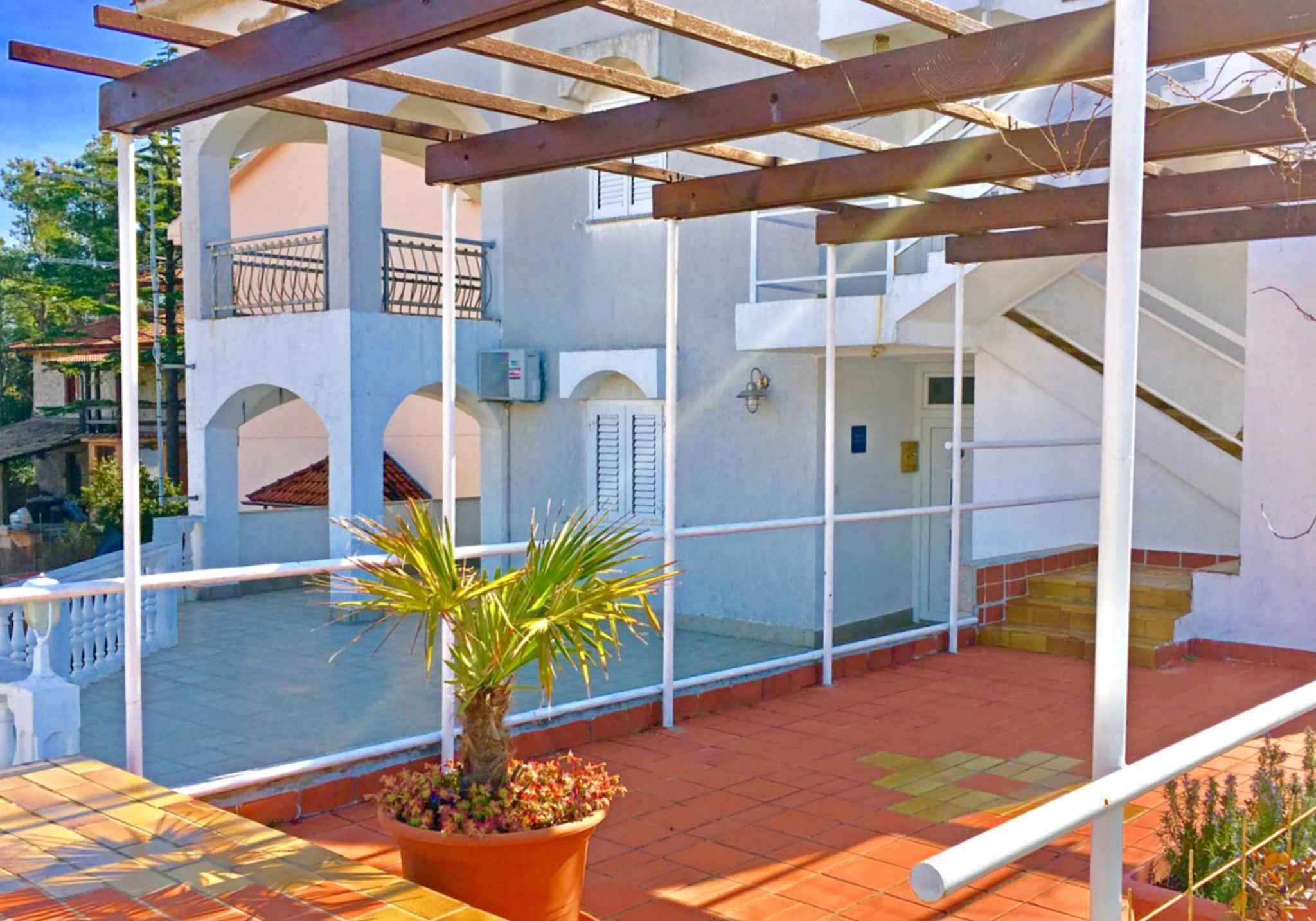 A covered outdoor area with a potted plant, a tiled floor, and a staircase leading to an upper level.