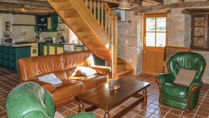 Un salon chaleureux avec un canapé en cuir marron, un fauteuil vert et une table basse en bois. Un escalier en bois mène à une cuisine dotée de meubles turquoise et d’un mur en pierre.