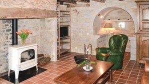 Une pièce rustique avec un mur en pierre, une table en bois, un fauteuil en cuir vert, un poêle blanc et un sol carrelé.