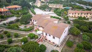 An aerial view of a coastal residential area with houses, a central building with a tiled roof, and a view of the sea and mountains in the background.