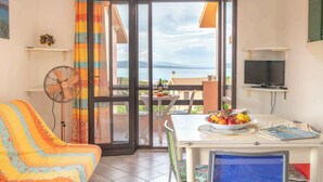 A room with a sofa, a table with fruit, a fan, a TV, and a view of the sea through a glass door.