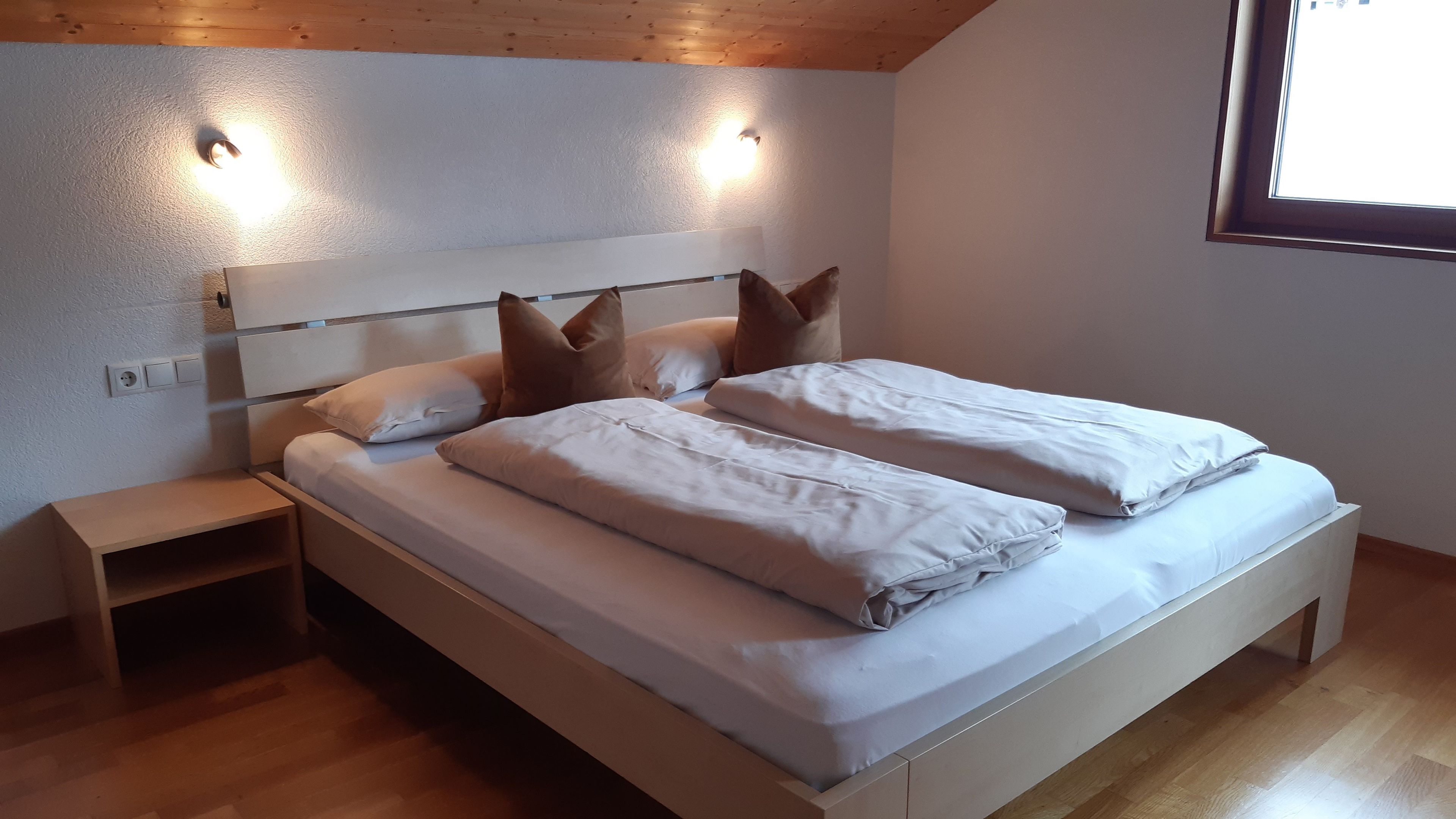 Ein Schlafzimmer mit einem hölzernen Bett, zwei Nachttischen, einem Kopfteil mit Wandleuchten und einem Fenster.