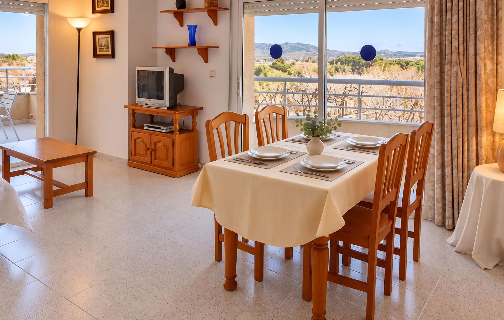 A dining area with a table set for four, chairs, a television on a stand, and a view of a desert landscape through large windows.