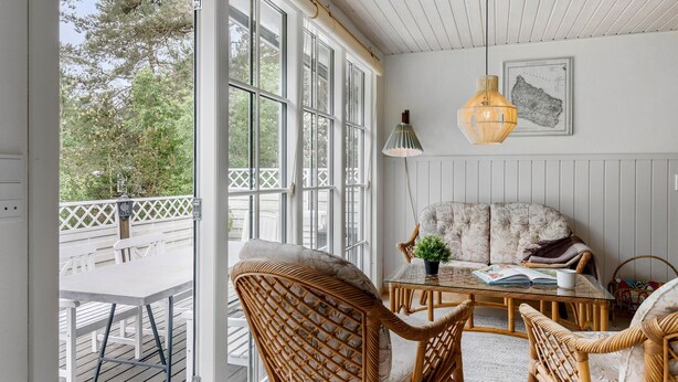 Una habitación soleada con ventanas amplias, un sofá de mimbre, una mesa de centro de vidrio y una lámpara colgante.