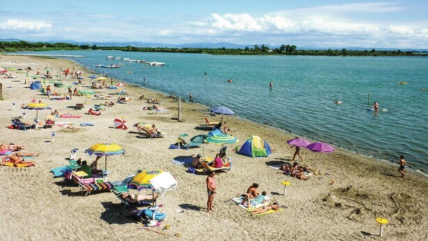 Ein Strand mit vielen Sonnenschirmen und Liegestühlen, ruhigem Meer und Booten in der Ferne.