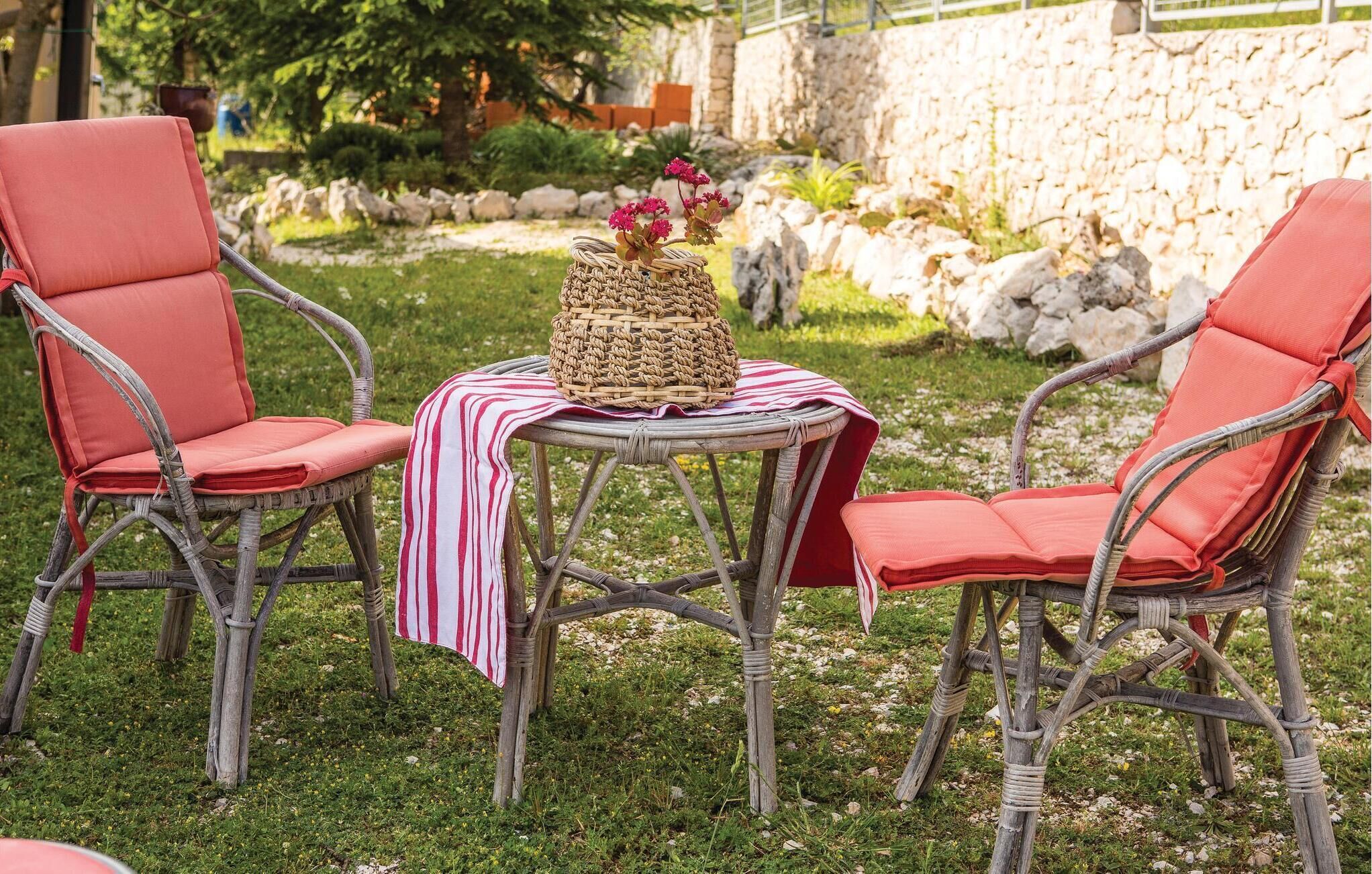 Due sedie in vimini con cuscini rossi, un cesto intrecciato e un panno a righe su un tavolino rotondo in un giardino.