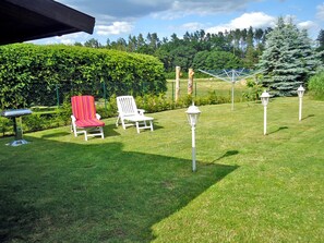 A grassy outdoor area with two white chairs, one with a red cushion, and a metal table. There are white lamp posts and a metal fence in the background. - Holiday house on Klostergrund - holiday home (Malchow)