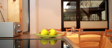 A modern kitchen with a glass table reflecting two apples on a green plate, wooden cabinets, and a window with a view of a house.