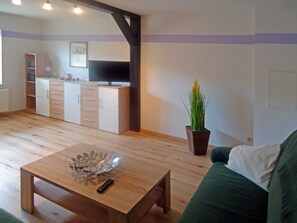 Ein Wohnzimmer mit Couchtisch aus Holz, einem grünen Sofa, einer Küchenzeile mit Schränken und einem Blumentopf.