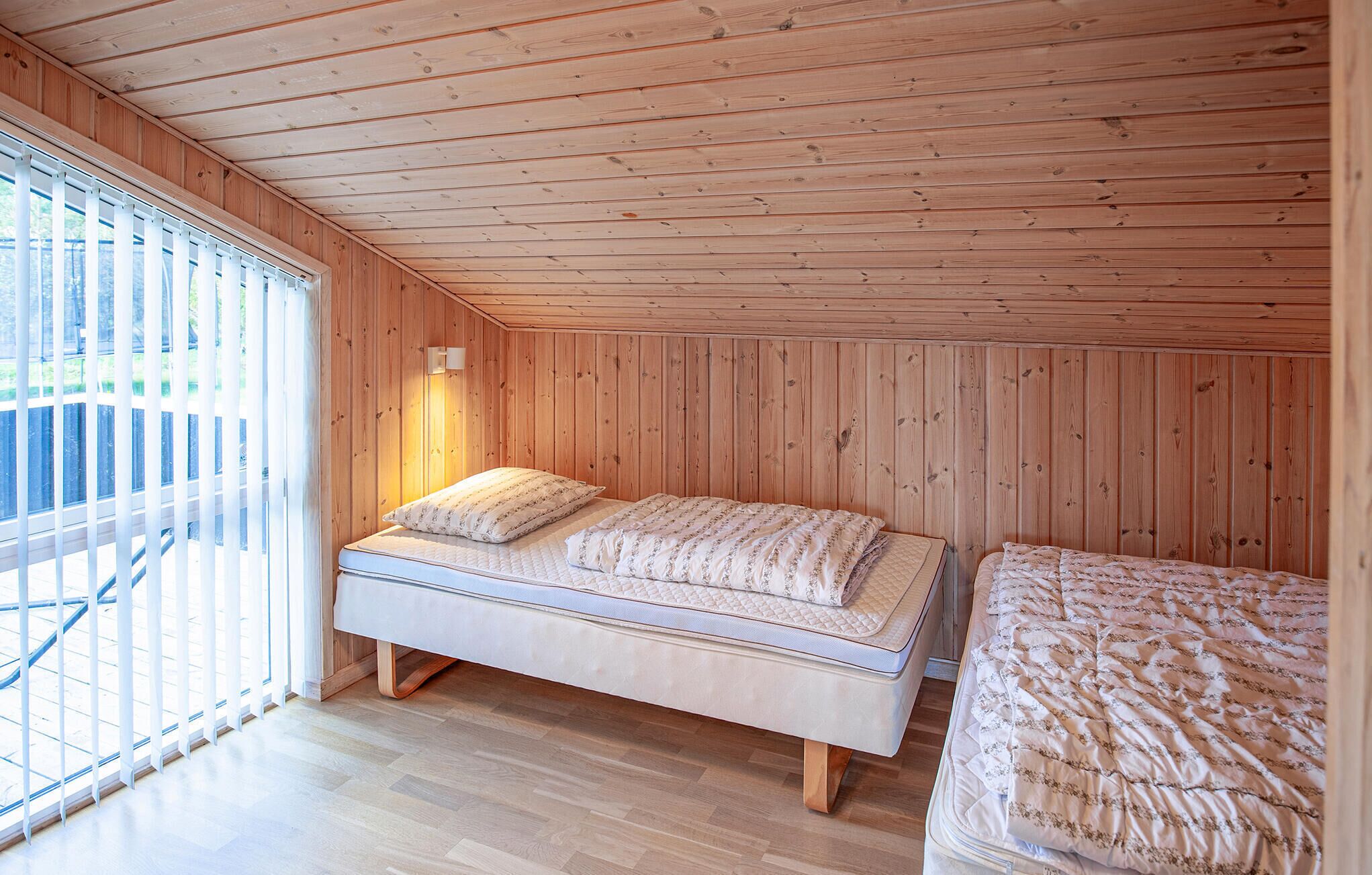 Un dormitorio con techo inclinado de madera, una cama con colchón acolchado y una ventana con persianas.