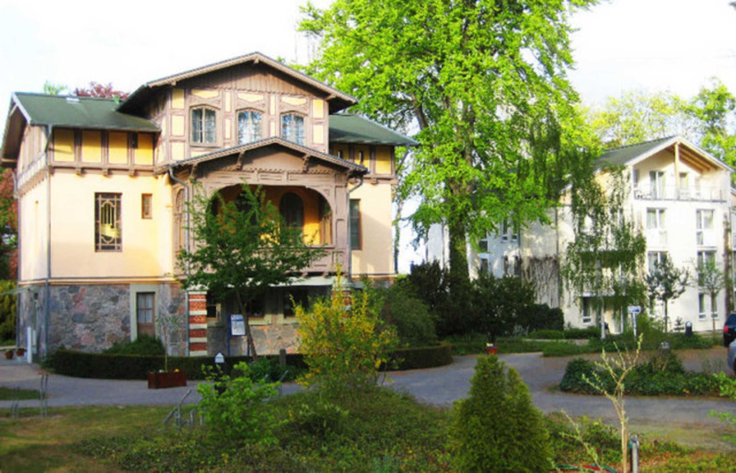 Un ampio edificio storico con un ingresso centrale, circondato da giardini curati e un'area di parcheggio.