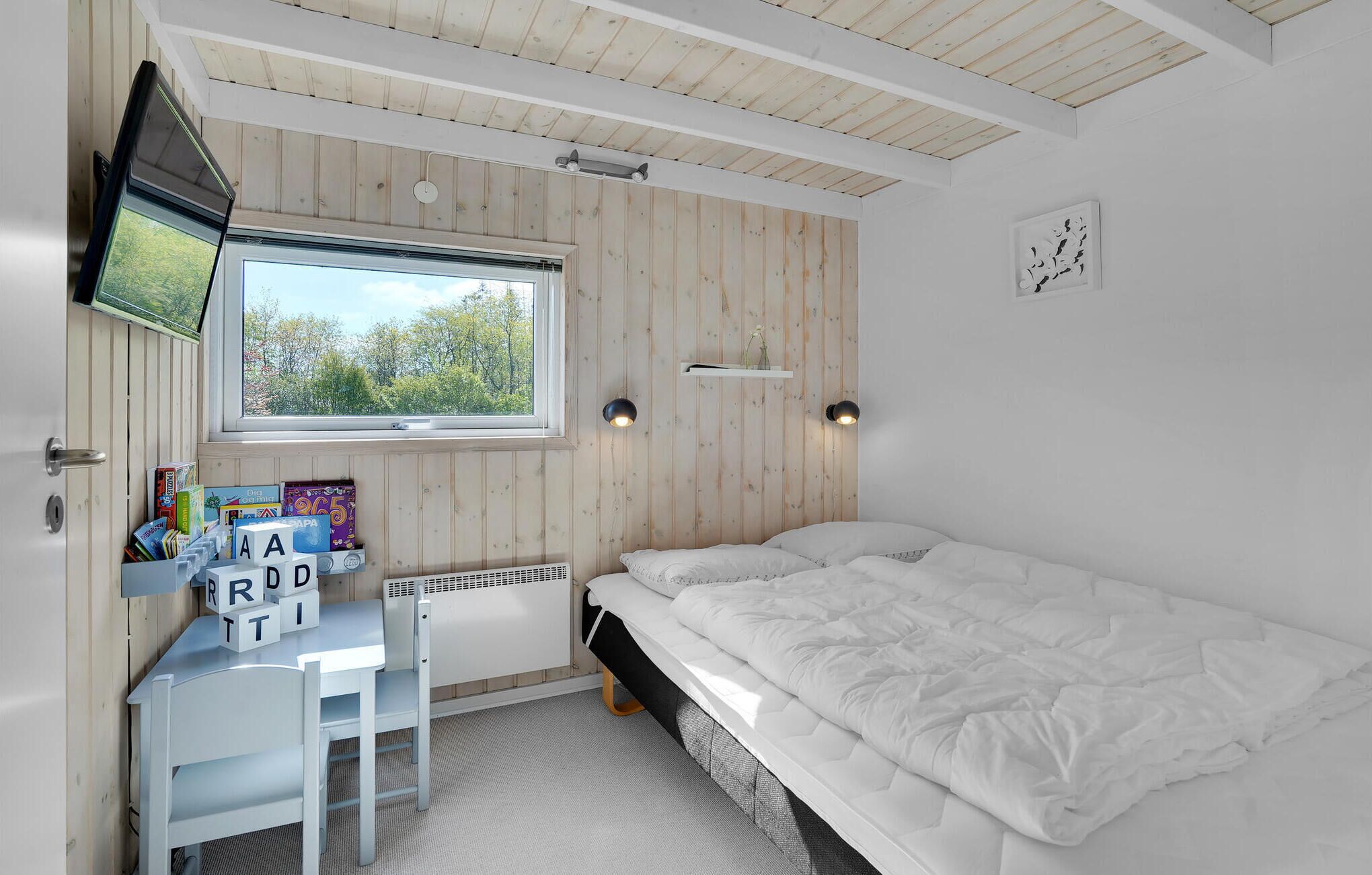 Un dormitorio con cama, un escritorio con juguetes educativos, una ventana con vistas a árboles y un televisor montado.