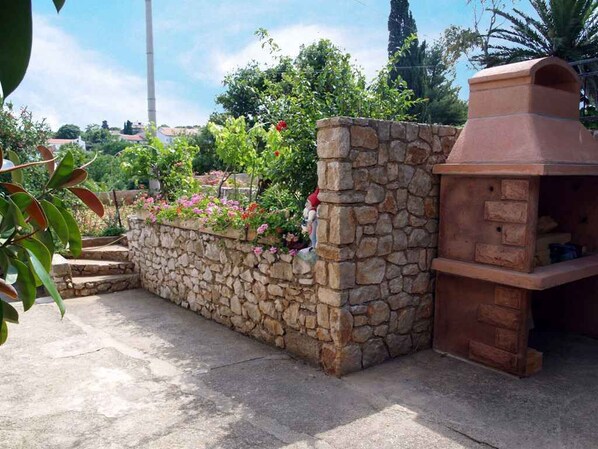 A stone wall with a built-in barbecue grill, surrounded by a garden with flowering plants.