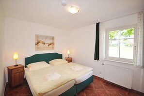 A bedroom with a bed, two bedside tables with lamps, a window with curtains, a radiator, and a painting on the wall.
