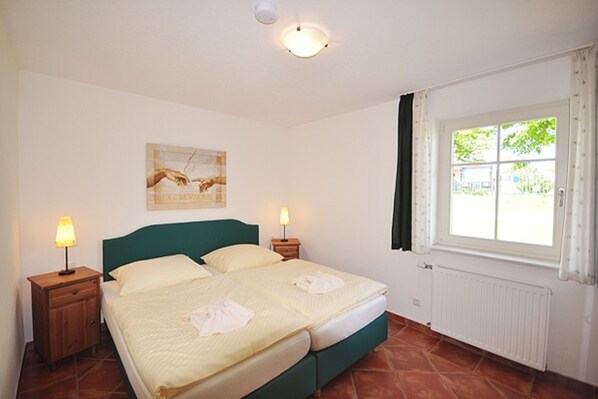 A bedroom with a bed, two bedside tables with lamps, a window with curtains, a radiator, and a painting on the wall. - 01 Holiday apartment with terrace - Karola's country house under the thatched roof (Mönchgut)