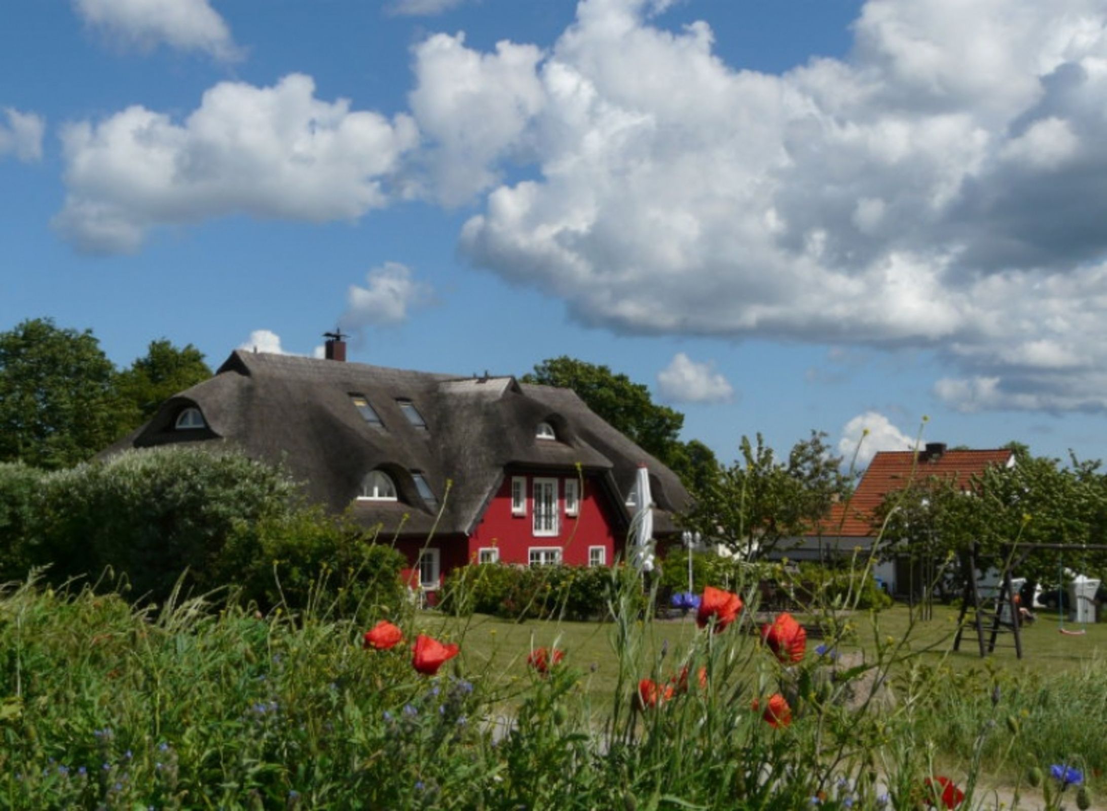 Une maison rouge avec un toit de chaume, entourée de verdure et de fleurs.