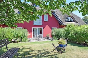 A red house with a thatched roof, a garden with a bench, and a wheelbarrow with plants.