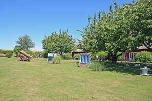 A grassy outdoor area with a wooden cart, a covered seating area, and a red building in the background. - 01 Holiday apartment with terrace - Karola's country house under the thatched roof (Mönchgut)