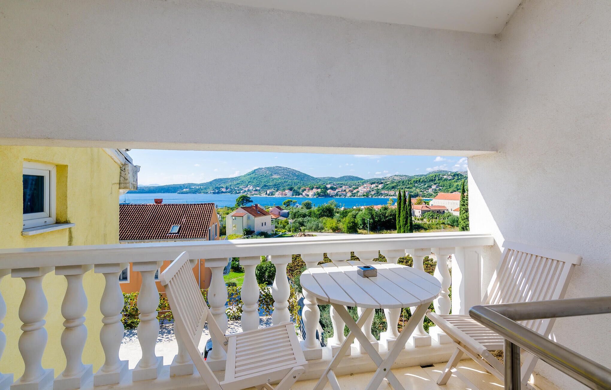 Un balcon doté d’une balustrade blanche, d’une petite table et d’une chaise. Le balcon offre une vue sur une ville côtière, avec des bâtiments et une montagne à l’horizon.