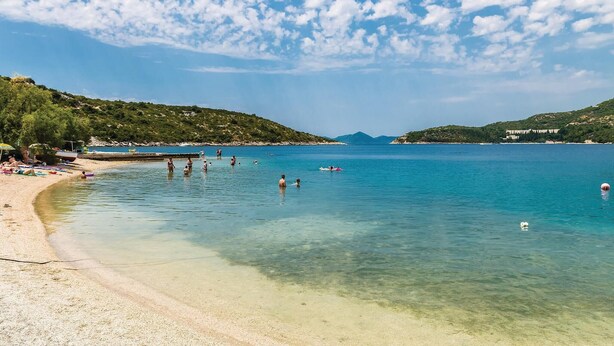 Una playa con agua cristalina, gente nadando y un paisaje montañoso al fondo.