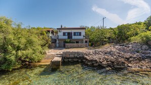 Une maison à deux étages avec un pont de pierre enjambant un ruisseau aux eaux claires, le tout entouré de verdure.