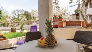 Une table sur laquelle est disposée une assiette de fruits, des chaises en osier et, en arrière-plan, une vue sur un jardin avec des arbres et un bâtiment.