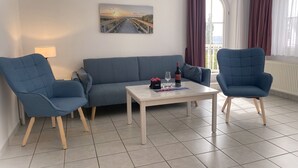 Ein modernes Wohnzimmer mit einer Couch, zwei Sesseln, einem Couchtisch mit einer Weinflasche, Gläsern und einem kleinen Blumenstrauß.