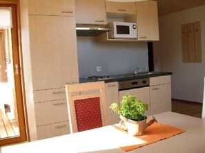 Cocina compacta con microondas, estufa y lavamanos. Hay una silla con cojín rojo y una planta en maceta sobre la encimera.