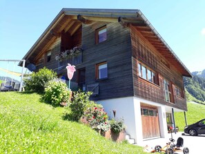 A two-story wooden house with a sloped roof, a satellite dish, and a car parked in front.